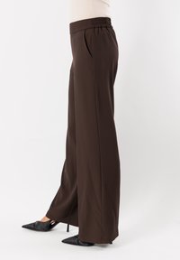 Pantaloni larghi marroni con vita smock, tasche laterali e una texture liscia, abbinati a tacchi neri appuntiti.