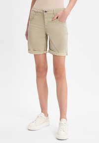 Khaki džíny šortky s ohrnutými manžetami, mají přední zapínání na knoflík, boční kapsy a hladkou texturu. V kombinaci s bílými teniskami.