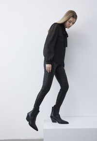 Zwarte doorzichtige blouse met gerimpelde halslijn, gecombineerd met zwarte gestreepte skinny jeans en puntige enkelbotten met een blokhak.
