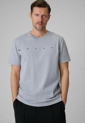 Mężczyzna noszący jasnoszarą koszulkę z krótkim rękawem z napisem "4F SPORT", stoi z rękami w kieszeniach czarnych spodni na jednolitym tle.