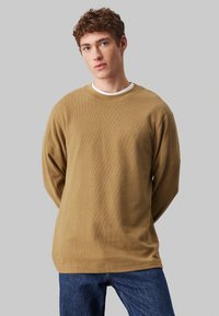 Sudadera marrón texturizada con cuello redondo y mangas largas, que presenta un logo bordado en el pecho, combinada con jeans de denim azul.