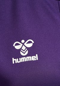Lila atletisk tyg med en framträdande vit Hummel-logotyp som har en stiliserad bi och geometriska designelement. Slät textur.
