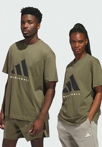 T-shirts desportivas verde azeitona com gráficos pretos, de corte descontraído e decote redondo, usadas com calções a condizer e bottoms cinzentos claros.