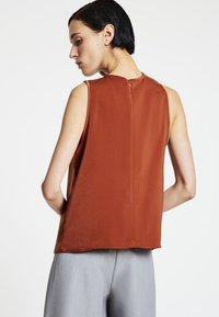 Top smanicato color ruggine con una texture liscia, caratterizzato da una cerniera posteriore e una vestibilità rilassata e squadrata. Abbinato a pantaloni grigio chiaro.