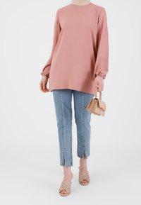 Różowa bluzka z długim rękawem i marszczonymi mankietami, niebieskie jeansy z krótkimi nogawkami i rozcięciami u dołu, cieliste sandały na obcasie oraz mała beżowa torebka.