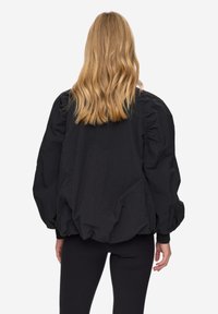 Černá, převeliká bunda s texturovanou látkou, objemnými rukávy a nazbieraným detailem na zadní straně, nošená přes přiléhavé černé legíny.