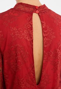 Rode kanten jurk met een textuurpatroon, voorzien van een sleutelgat achterkant en twee rode knoopsluitingen bij de hals. Glad stoffen afwerking.