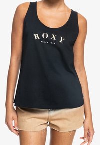 Czarny bawełniany top na ramiączkach z okrągłym dekoltem oraz napisem "ROXY" w złotym nadruku, zestawiony z jasnobrązowymi szortami dżinsowymi z postrzępionymi brzegami.