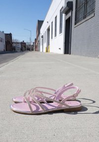 Paire de sandales roses à brides posées sur un trottoir urbain ensoleillé avec des bâtiments blancs et gris en arrière-plan.
