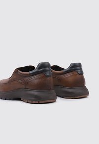 Zapatos de cuero marrón con un acabado texturizado, acentos en el talón negro y una suela de goma con patrones de textura visibles. Vista trasera mostrada.