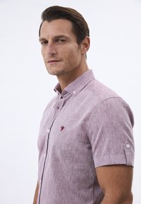 Kurzärmliges, hellrosa Button-up-Shirt aus strukturiertem Stoff, mit einem kleinen gestickten Vogel-Logo auf der Brust und weißen Knöpfen.