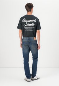 Camiseta negra con texto en cursiva blanca en la parte posterior, combinada con jeans de mezclilla azul medio y calzado gris. Diseño simple, ajuste casual.