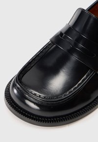 Nærbilde av en blank svart skinnloafersko med firkantet tå og dekorativ stropp på hvit bakgrunn.