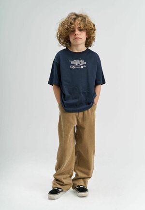 Tengerész kék pamut póló grafikai nyomattal, bézs kordbársony bővített nadrággal párosítva. Fekete sportcipők fehér talppal. A modell keze a zsebében áll.