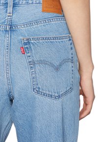 Jeans de mezclilla azul claro con un diseño de bolsillo trasero curvado, etiqueta de logo roja y parche de cuero marrón en la cintura. Detalle de costura visible.