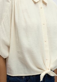 Chemise texturée crème à manches courtes, boutonnée et nouée à l'ourlet, portée avec un jean en denim bleu sur une personne.