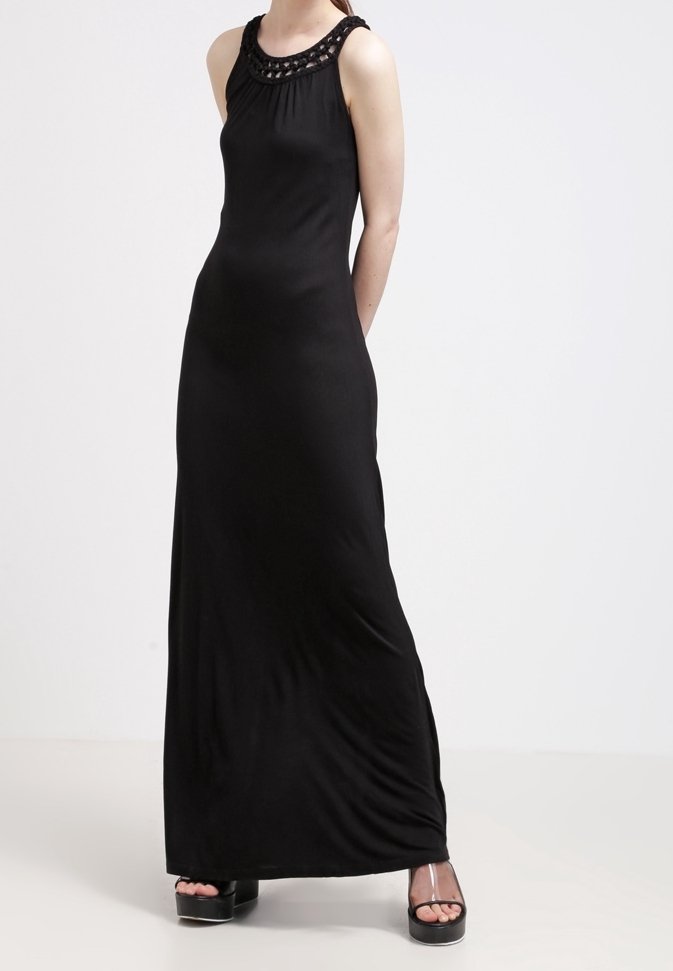 Femme portant une robe maxi noire sans manches avec un col tressé, associée à des sandales à plateforme noires avec des brides transparentes.