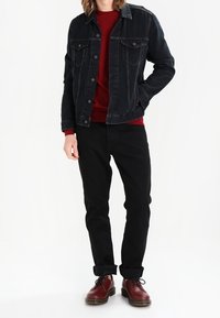 Zwarte denimjack over een rode trui, gecombineerd met zwarte broek en bordeauxrode schoenen. Kenmerken zijn onder andere een knoopsluiting en opgerolde broekspijpen.