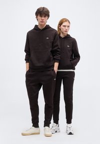 Νεαρός άνδρας και γυναίκα στέκονται δίπλα-δίπλα φορώντας μαύρα φούτερ με κουκούλα και παντελόνια φόρμας ταιριαστά, με λευκά αθλητικά παπούτσια, ποζάροντας μπροστά σε απλό φόντο.