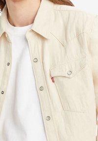 Vaaleanbeige nappipaita teksturoidusta kankaasta, jossa on rintatasku ja hopeiset painonapit. Päällä valkoinen t-paita.