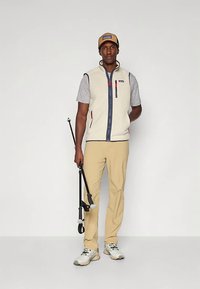 Άνδρας φοράει μπεζ γιλέκο, γκρι μπλουζάκι, μπεζ παντελόνι και καπέλο, κρατώντας διπλωμένα μαύρα κοντάρια πεζοπορίας σε απλή λευκή φόντο.