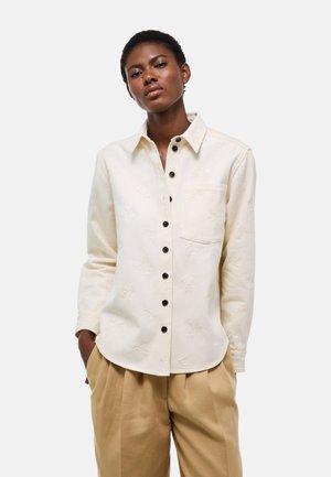 Femme noire portant une chemise crème boutonnée avec un motif floral texturé et un pantalon plissé beige, debout les mains dans les poches.