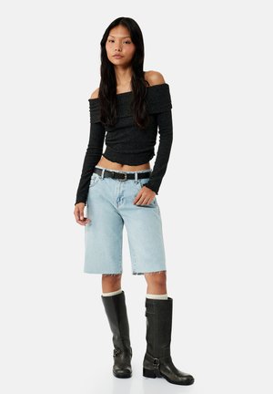 Jonge vrouw die een zwarte off-shoulder top met lange mouwen draagt, lichtblauwe denim shorts tot op de knie, een riem en zwarte kniehoge laarzen met witte sokken.