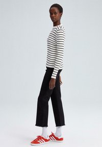 Zwart-witte gestreepte ribbeltrui, zwarte cropped jeans met rafelige rand en rode sneakers met witte strepen. Model staat zijwaarts.