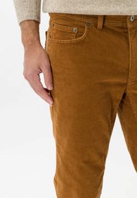 Bruna kordfläckiga byxor har en texturerad, ribbad yta med framfickor och metallspännen. Handen som vilar på låret visar passformen.