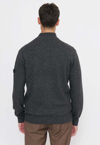 Uomo di spalle che indossa un maglione grigio scuro a collo alto e pantaloni marroni, su uno sfondo semplice.