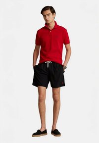 Sarkans polo krekls ar apkakli, melnas šorti ar elastīgu jostu un auklu, un melnas slip-on kurpes ar jutas zolēm.