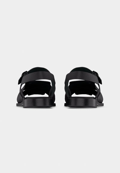 Paire de sandales noires à bout ouvert avec brides réglables à la cheville et semelles plates, vues de dos sur un fond uni.
