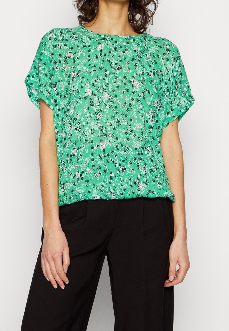 Blusa floral verde de manga corta, con una tela ligera y un corte holgado. Presenta patrones florales en blanco y negro. Combinada con pantalones negros.