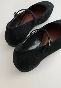 Ballerines en daim noir avec un bout rond, une sangle à la cheville et un petit détail de boucle. Intérieur lisse avec une semelle plate en caoutchouc.