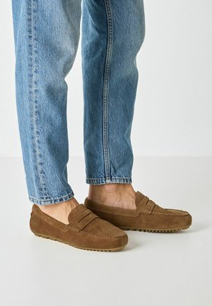 Persona che indossa jeans azzurri e mocassini in suede marrone, in piedi su un pavimento chiaro.
