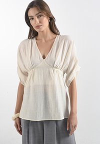 Donna con lunghi capelli che indossa una camicia bianca ampia e una gonna grigia, in piedi su uno sfondo bianco, che guarda verso la macchina fotografica.