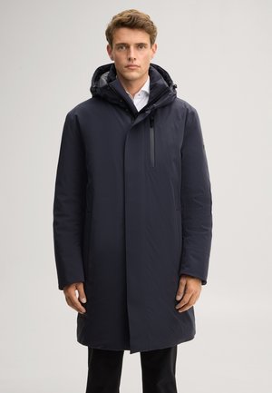 Man die een lange marineblauwe winterjas met kap draagt over een wit overhemd, staande tegen een effen lichte achtergrond.