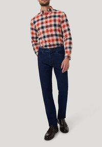 Geruite flanellen shirt in rood, blauw en crème; donkerblauwe jeans; zwarte loafers. Shirt heeft een button-down kraag en een borstzak.