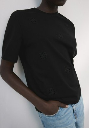 Persoană purtând o cămașă neagră cu mâneci scurte, cu detalii subtile decupate în formă de flori și blugi albastru deschis, stând cu o mână în buzunar.