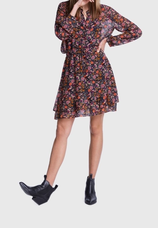 Femme portant une robe longue à manches longues à motif floral foncé avec une ceinture à la taille et des bottines noires, posant avec une jambe tendue et la main près du visage.
