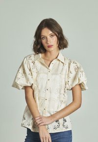 Cremige Spitzenbluse mit Puffärmeln, Knopfverschluss und Blumenmuster; verfügt über einen strukturierten Stoff und einen subtilen Glanz. Kombiniert mit blauer Jeans.