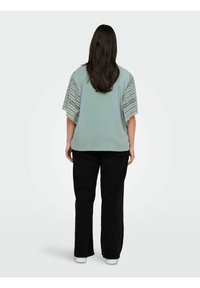Top verde chiaro con dettagli in pizzo sulle spalle, maniche fluttuanti corte e vestibilità rilassata, abbinato a pantaloni neri a gamba larga.