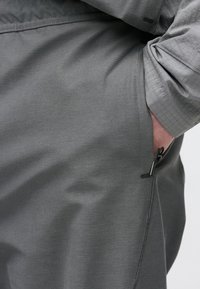 Γκρι παντελόνι με λεία υφή, που διαθέτει πλαϊνή τσέπη με φερμουάρ και μερικώς γυρισμένο μανίκι ενός ταιριαστού γκρι τοπ.