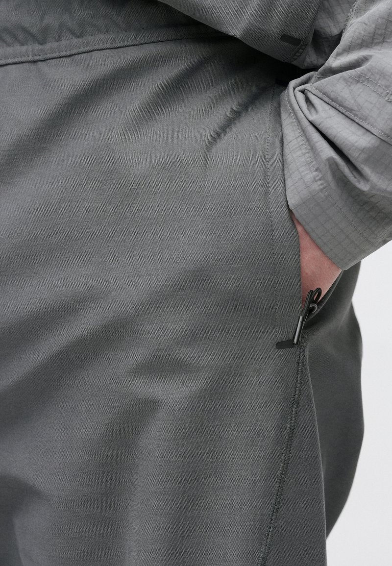 Grå bukser med en glat tekstur, med en sidelomme med lynlås og en delvist rullet ærme på en matchende grå top.