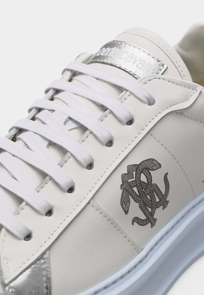 Λευκό δερμάτινο αθλητικό παπούτσι με λευκά κορδόνια, ασημί ταμπελάκι στη φτέρνα, ασημί ετικέτα μάρκας και μαύρο λογότυπο φιδιού στην πλαϊνή επιφάνεια.