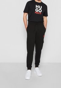 Zwarte katoenen t-shirt met vetgedrukte witte en rode tekst met de opdruk "HUGO FOR IT." Gecombineerd met zwarte joggers met een zijzak en trekkoord. Witte sneakers.
