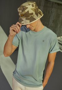 T-shirt en coton bleu clair avec des manches roulées, associée à un chapeau seau camouflage beige et marron avec un patch logo. Fond en béton.