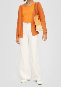 Oranger Cardigan über einem orangen Oberteil, kombiniert mit weißen, weit geschnittenen Hosen. Accessoire: eine beige-weiße Bucket-Tasche. Weiße Sneakers.
