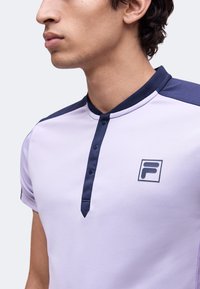 Jeune homme portant une chemise violet clair avec des boutons bleu marine foncé et des accents aux épaules, montrant un logo rectangulaire avec un "F" sur la poitrine.