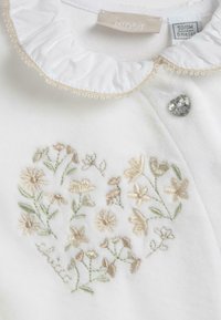 Vêtement en tissu blanc avec un col volanté, présentant une broderie florale dans des tons de beige doux et de vert, ainsi qu'un bijou accent en forme de cœur.